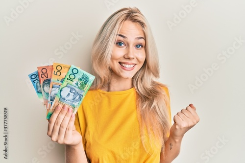 Beautiful caucasian blonde girl holding australian dollars screaming proud, celebrating victory and success very excited with raised arm