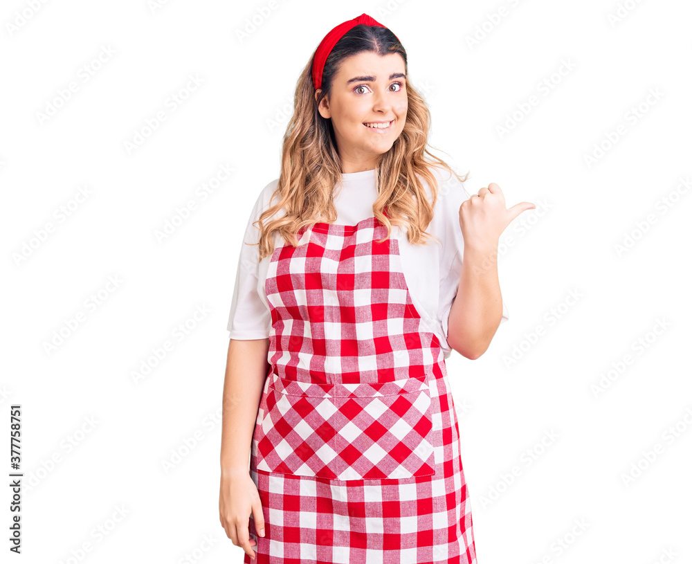 Young caucasian woman wearing apron smiling with happy face looking and pointing to the side with thumb up. Young caucasian woman wearing apron smiling with happy face looking and pointing to the side with thumb up.