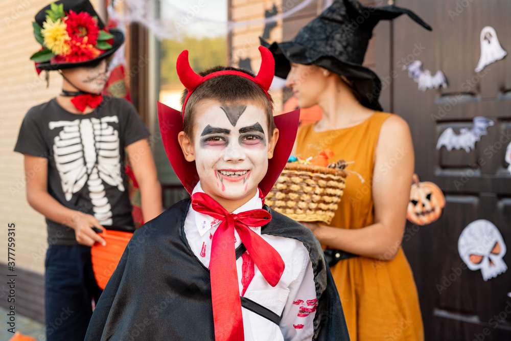Cheerful boy in halloween costume looking at you against young woman and friend