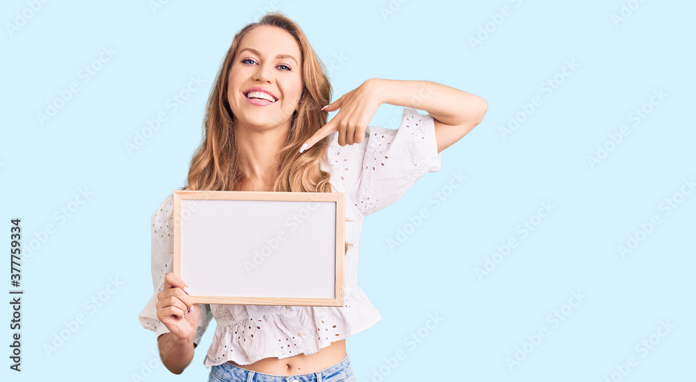 Young caucasian woman with blond hair holding blank empty banner pointing finger to one self smiling happy and proud Young caucasian woman with blond hair holding blank empty banner pointing finger to one self smiling happy and proud