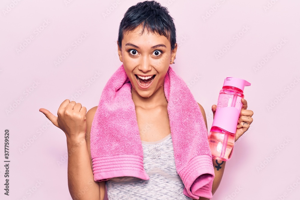 Young woman wearing sportswearholding water bottle pointing thumb up to the side smiling happy with open mouth Young woman wearing sportswearholding water bottle pointing thumb up to the side smiling happy with open mouth