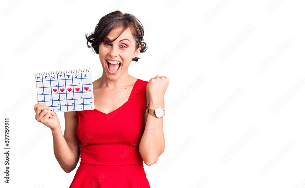 Beautiful young woman with short hair holding heart calendar screaming proud, celebrating victory and success very excited with raised arms Beautiful young woman with short hair holding heart calendar screaming proud, celebrating victory and success very excited with raised arms