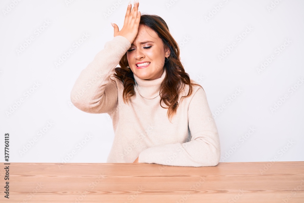 Young beautiful caucasian woman wearing turtleneck sweater sitting on the table surprised with hand on head for mistake, remember error. forgot, bad memory concept. Young beautiful caucasian woman wearing turtleneck sweater sitting on the table surprised with hand on head for mistake, remember error. forgot, bad memory concept.