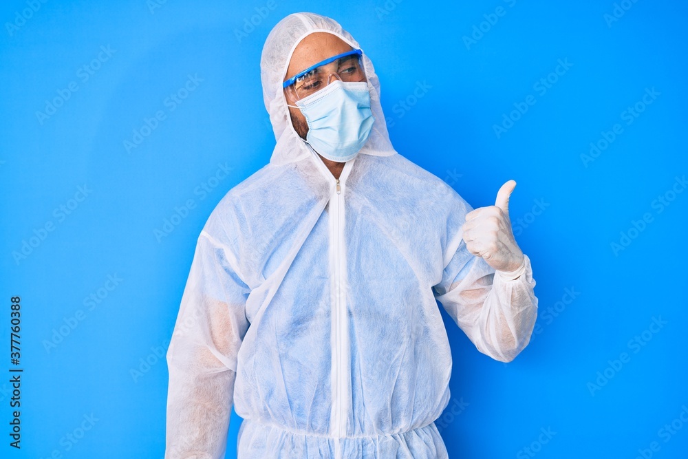 Young hispanic man wearing doctor coronavirus protection uniform smiling with happy face looking and pointing to the side with thumb up. Young hispanic man wearing doctor coronavirus protection uniform smiling with happy face looking and pointing to the side with thumb up.