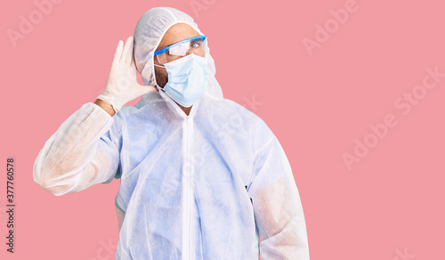 Young hispanic man wearing doctor protection coronavirus uniform and medical mask smiling with hand over ear listening an hearing to rumor or gossip. deafness concept. photo