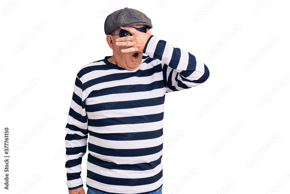 Senior handsome man wearing burglar mask and t-shirt peeking in shock covering face and eyes with hand, looking through fingers with embarrassed expression. Senior handsome man wearing burglar mask and t-shirt peeking in shock covering face and eyes with hand, looking through fingers with embarrassed expression.
