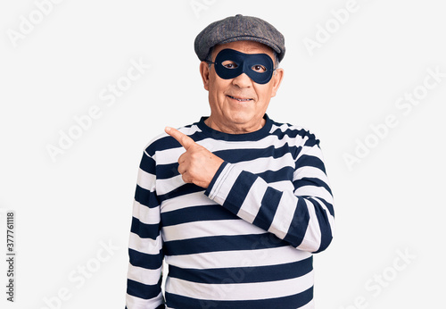 Senior handsome man wearing burglar mask and t-shirt cheerful with a smile on face pointing with hand and finger up to the side with happy and natural expression