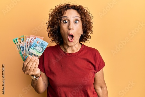 Beautiful middle age mature woman holding australian dollars scared and amazed with open mouth for surprise, disbelief face