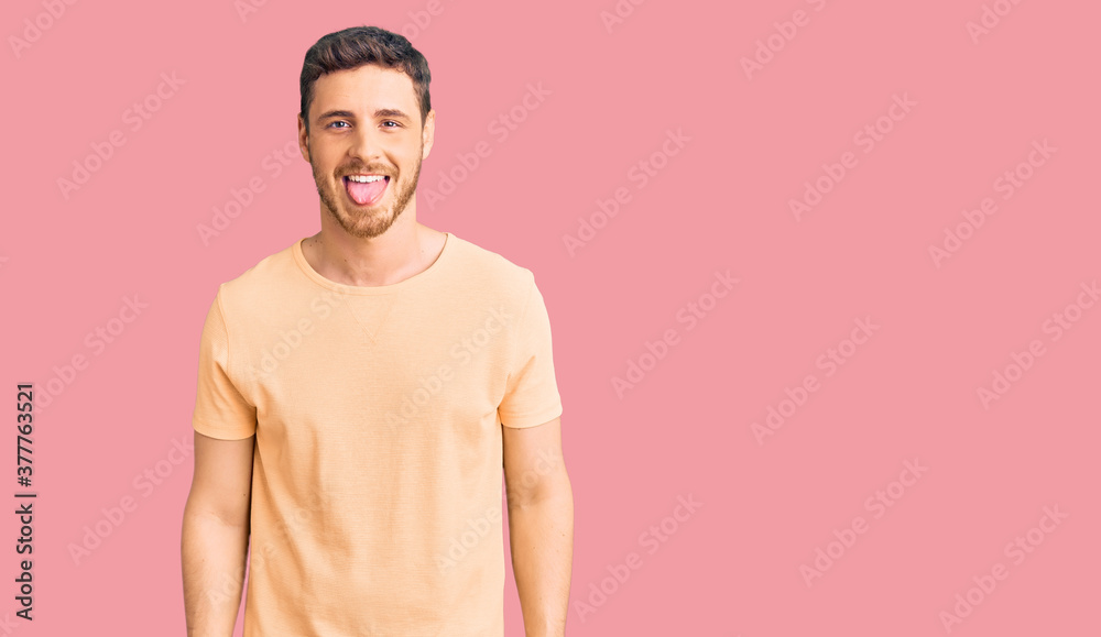 Handsome young man with bear wearing casual yellow tshirt sticking tongue out happy with funny expression. emotion concept.