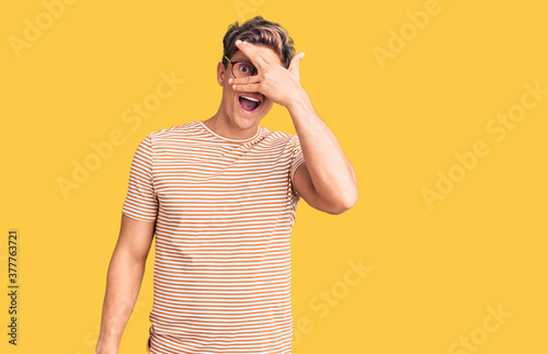 Young handsome man wearing casual clothes and glasses peeking in shock covering face and eyes with hand, looking through fingers with embarrassed expression.