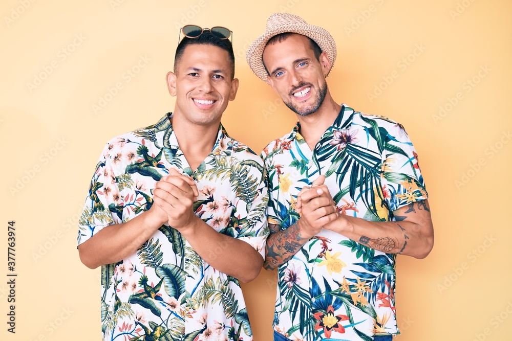 Young gay couple of two men wearing summer hat and hawaiian shirt with hands together and crossed fingers smiling relaxed and cheerful. success and optimistic Young gay couple of two men wearing summer hat and hawaiian shirt with hands together and crossed fingers smiling relaxed and cheerful. success and optimistic