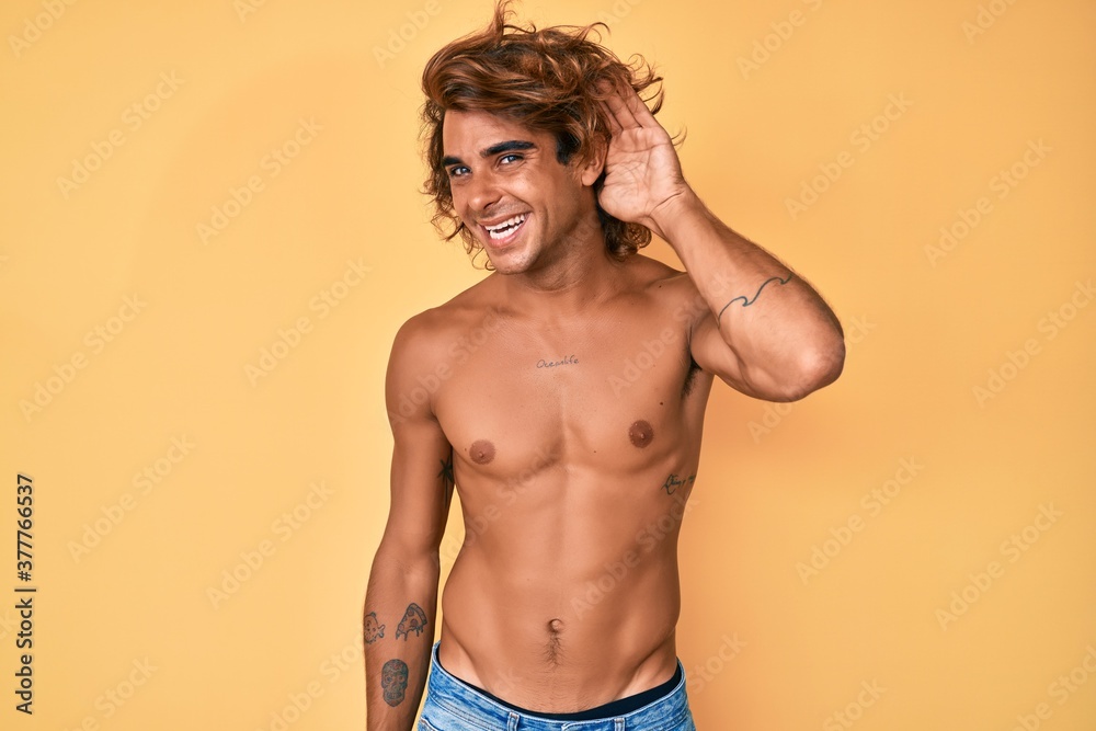 Fototapeta premium Young hispanic man standing shirtless smiling with hand over ear listening an hearing to rumor or gossip. deafness concept.