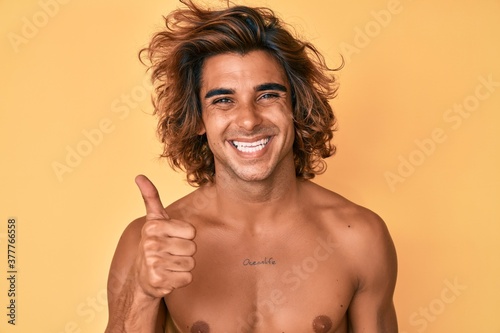 Young hispanic man standing shirtless smiling happy and positive  thumb up doing excellent and approval sign