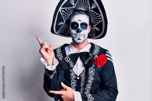 Young man wearing mexican day of the dead costume over white with a big smile on face, pointing with hand and finger to the side looking at the camera.