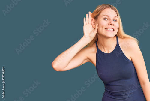 Young beautiful blonde woman wearing casual dress smiling with hand over ear listening an hearing to rumor or gossip. deafness concept.