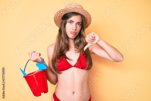 Beautiful young caucasian woman playing with summer shovel and bucket toys with angry face, negative sign showing dislike with thumbs down, rejection concept