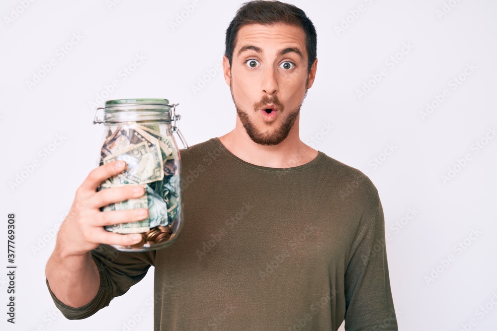 Young handsome man holding jar with savings scared and amazed with open mouth for surprise, disbelief face Young handsome man holding jar with savings scared and amazed with open mouth for surprise, disbelief face