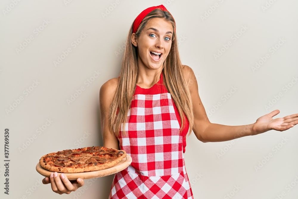 Young blonde woman wearing professional baker apron holding italian pizza celebrating achievement with happy smile and winner expression with raised hand Young blonde woman wearing professional baker apron holding italian pizza celebrating achievement with happy smile and winner expression with raised hand