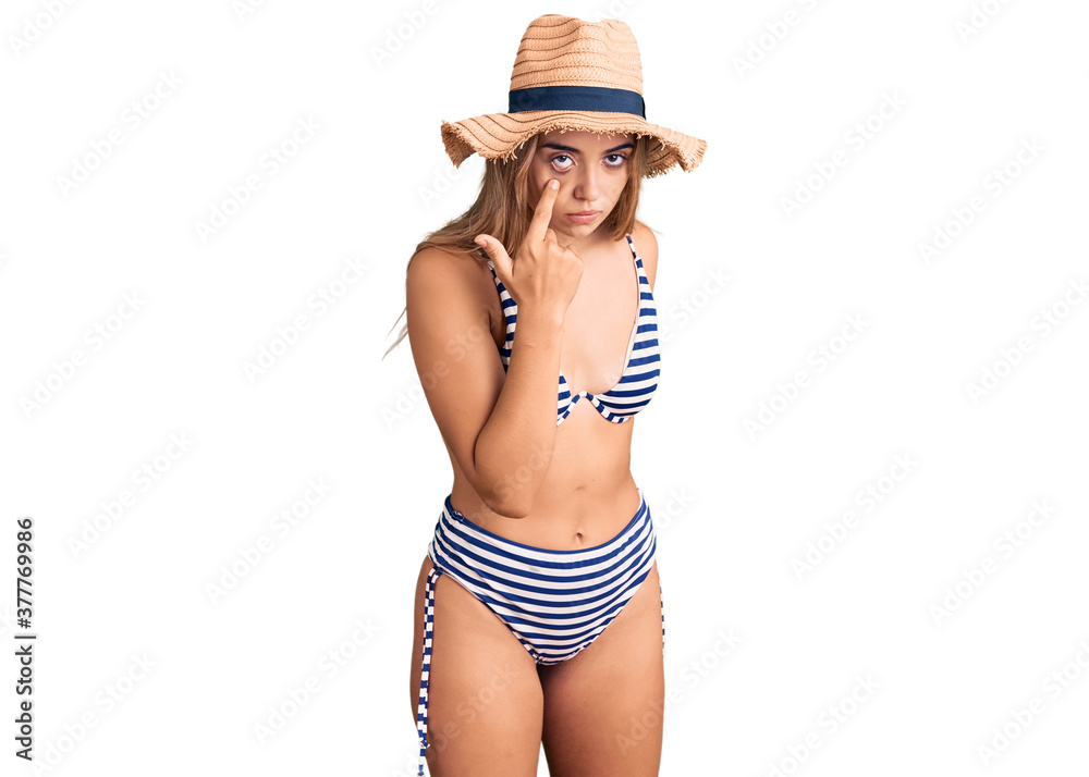 Young beautiful blonde woman wearing bikini and hat pointing to the eye watching you gesture, suspicious expression Young beautiful blonde woman wearing bikini and hat pointing to the eye watching you gesture, suspicious expression