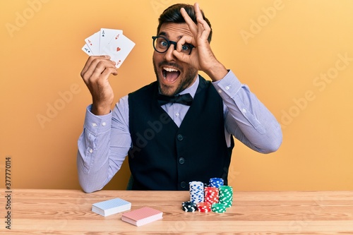 Handsome hispanic croupier man sitting on the table with poker chips and cards smiling happy doing ok sign with hand on eye looking through fingers