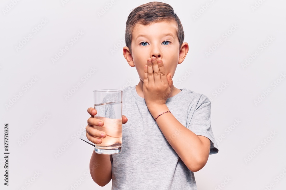 Cute blond kid drinking glass of water covering mouth with hand, shocked and afraid for mistake. surprised expression Cute blond kid drinking glass of water covering mouth with hand, shocked and afraid for mistake. surprised expression
