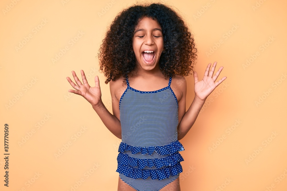 African american child with curly hair wearing swimwear celebrating mad and crazy for success with arms raised and closed eyes screaming excited. winner concept African american child with curly hair wearing swimwear celebrating mad and crazy for success with arms raised and closed eyes screaming excited. winner concept