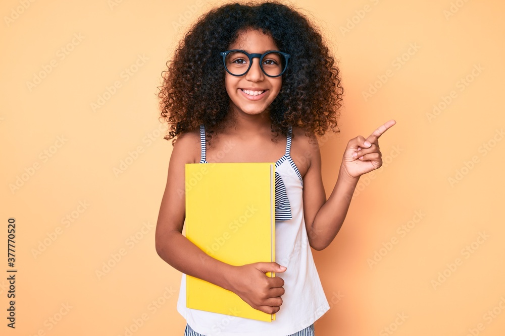 African american child with curly hair wearing glasses and holding book smiling happy pointing with hand and finger to the side African american child with curly hair wearing glasses and holding book smiling happy pointing with hand and finger to the side