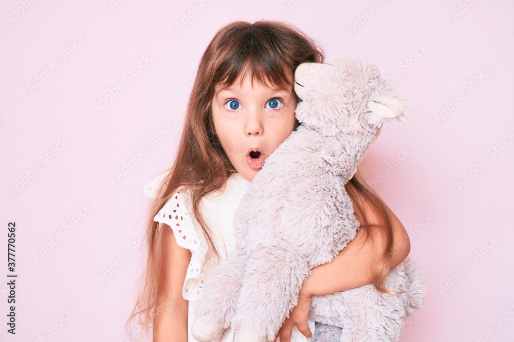 Little caucasian kid girl with long hair holding stuffed animal scared and amazed with open mouth for surprise, disbelief face Little caucasian kid girl with long hair holding stuffed animal scared and amazed with open mouth for surprise, disbelief face