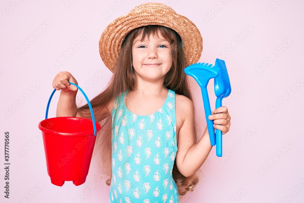 Little caucasian kid girl with long hair playing with summer shovel and bucket toys looking positive and happy standing and smiling with a confident smile showing teeth Little caucasian kid girl with long hair playing with summer shovel and bucket toys looking positive and happy standing and smiling with a confident smile showing teeth
