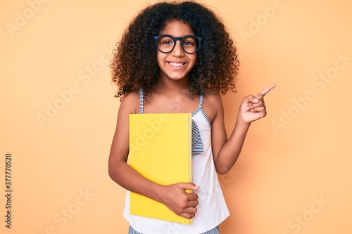 African american child with curly hair wearing glasses and holding book smiling happy pointing with hand and finger to the side