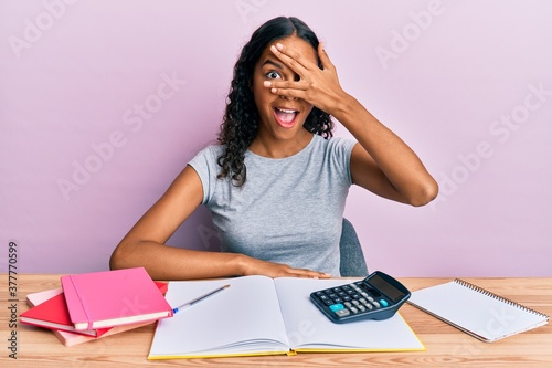 Young african american girl accountant working at the office peeking in shock covering face and eyes with hand, looking through fingers with embarrassed expression.
