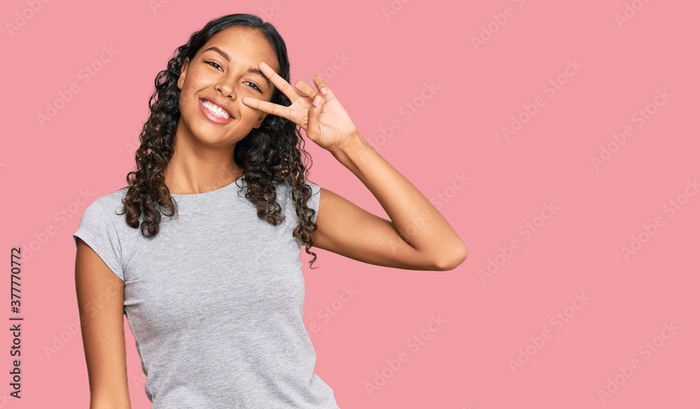 Fototapeta premium Young african american girl wearing casual clothes doing peace symbol with fingers over face, smiling cheerful showing victory