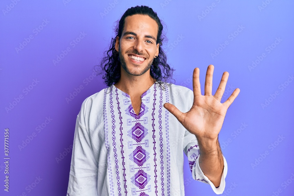 Young handsome man with long hair wearing bohemian and hippie shirt showing and pointing up with fingers number five while smiling confident and happy. Young handsome man with long hair wearing bohemian and hippie shirt showing and pointing up with fingers number five while smiling confident and happy.