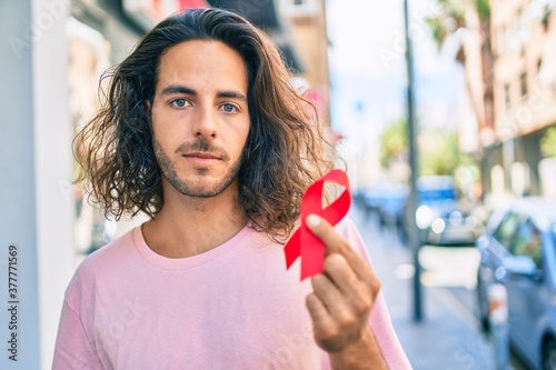 Картината върху платно Young hispanic man with serious expression holding hiv awareness red ribbon at city