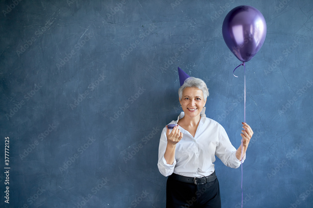 Elegant gray haired mature female wearing cone hat and stylish clothes eating macaroon at party, holding air balloon, posing against blank studio wall background with copy space for your advertisement Elegant gray haired mature female wearing cone hat and stylish clothes eating macaroon at party, holding air balloon, posing against blank studio wall background with copy space for your advertisement