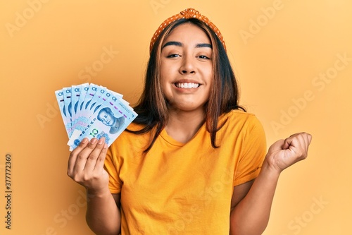 Young latin woman holding yuan chinese banknotes screaming proud, celebrating victory and success very excited with raised arm