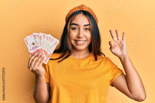 Young latin woman holding colombian pesos doing ok sign with fingers, smiling friendly gesturing excellent symbol