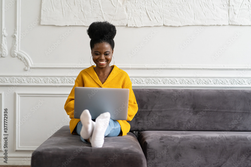 Smiling African American millennial woman with afro hairstyle wear yellow cardigan sitting on sofa, resting, looking at camera webcam and talking on a video call or skype with friends, watching movie - obrazy, fototapety, plakaty 