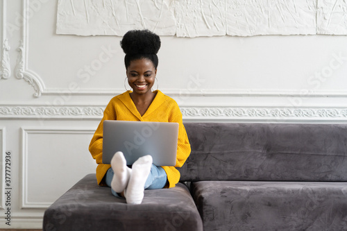 Smiling African American millennial woman with afro hairstyle wear yellow cardigan sitting on sofa, resting, looking at camera webcam and talking on a video call or skype with friends, watching movie photo