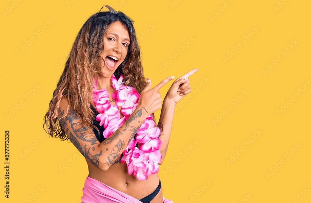 Young hispanic woman with tattoo wearing bikini and hawaiian lei smiling and looking at the camera pointing with two hands and fingers to the side.