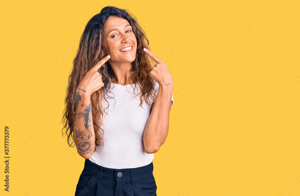 Young hispanic woman with tattoo wearing casual white tshirt smiling cheerful showing and pointing with fingers teeth and mouth. dental health concept. Young hispanic woman with tattoo wearing casual white tshirt smiling cheerful showing and pointing with fingers teeth and mouth. dental health concept.
