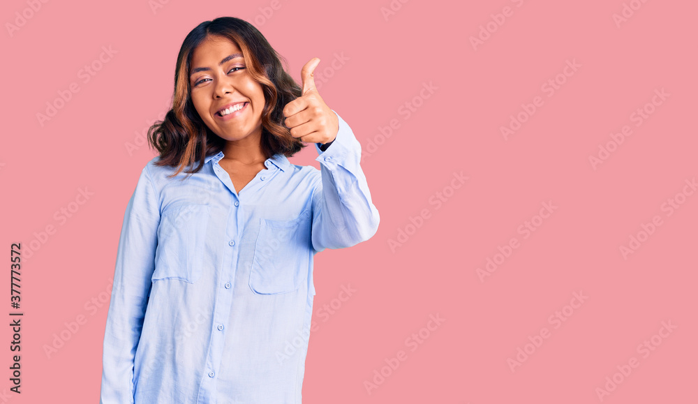 Young beautiful mixed race woman wearing casual business shirt smiling happy and positive, thumb up doing excellent and approval sign Young beautiful mixed race woman wearing casual business shirt smiling happy and positive, thumb up doing excellent and approval sign