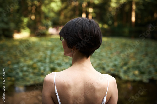 Outdoor rear view of attractive young brunette girl with short haircut admiring beautiful wild nature  spending weekend outside in park  standing in front of pond with water lilies in background