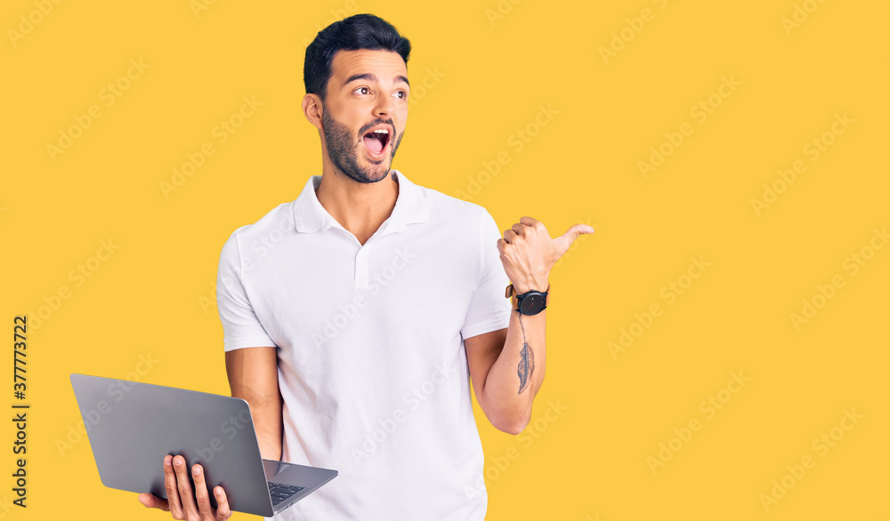Young handsome hispanic man working using computer laptop pointing thumb up to the side smiling happy with open mouth Young handsome hispanic man working using computer laptop pointing thumb up to the side smiling happy with open mouth