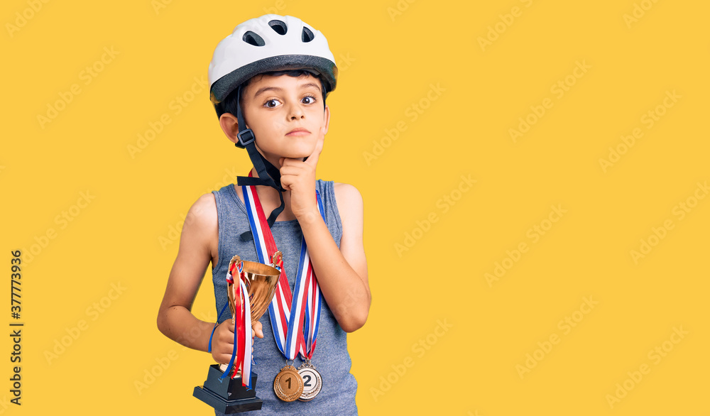 Little cute boy kid wearing bike helmet and winner medals holding winner trophy serious face thinking about question with hand on chin, thoughtful about confusing idea Little cute boy kid wearing bike helmet and winner medals holding winner trophy serious face thinking about question with hand on chin, thoughtful about confusing idea