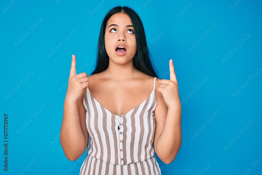Young beautiful asian girl wearing casual clothes amazed and surprised looking up and pointing with fingers and raised arms. Young beautiful asian girl wearing casual clothes amazed and surprised looking up and pointing with fingers and raised arms.