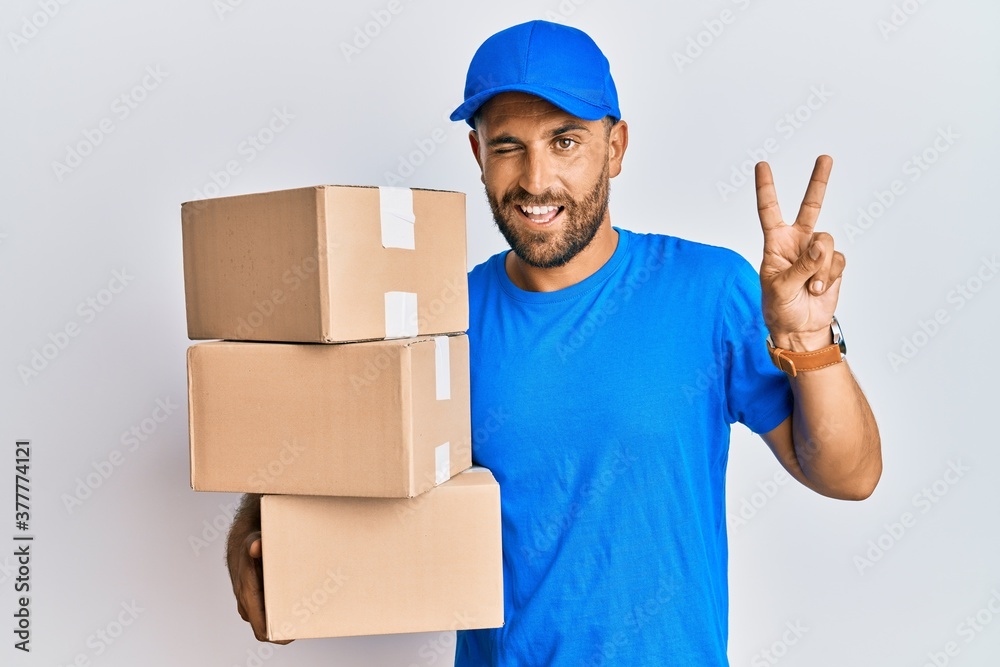 Handsome man with beard wearing courier uniform holding delivery packages smiling with happy face winking at the camera doing victory sign with fingers. number two. Handsome man with beard wearing courier uniform holding delivery packages smiling with happy face winking at the camera doing victory sign with fingers. number two.
