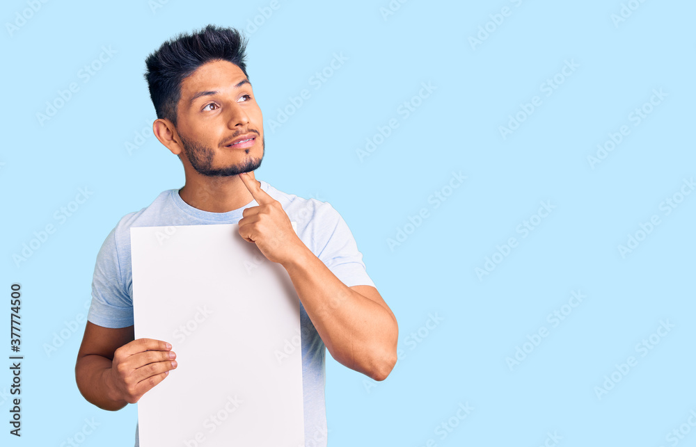 Handsome latin american young man holding cardboard banner with blank space serious face thinking about question with hand on chin, thoughtful about confusing idea Handsome latin american young man holding cardboard banner with blank space serious face thinking about question with hand on chin, thoughtful about confusing idea