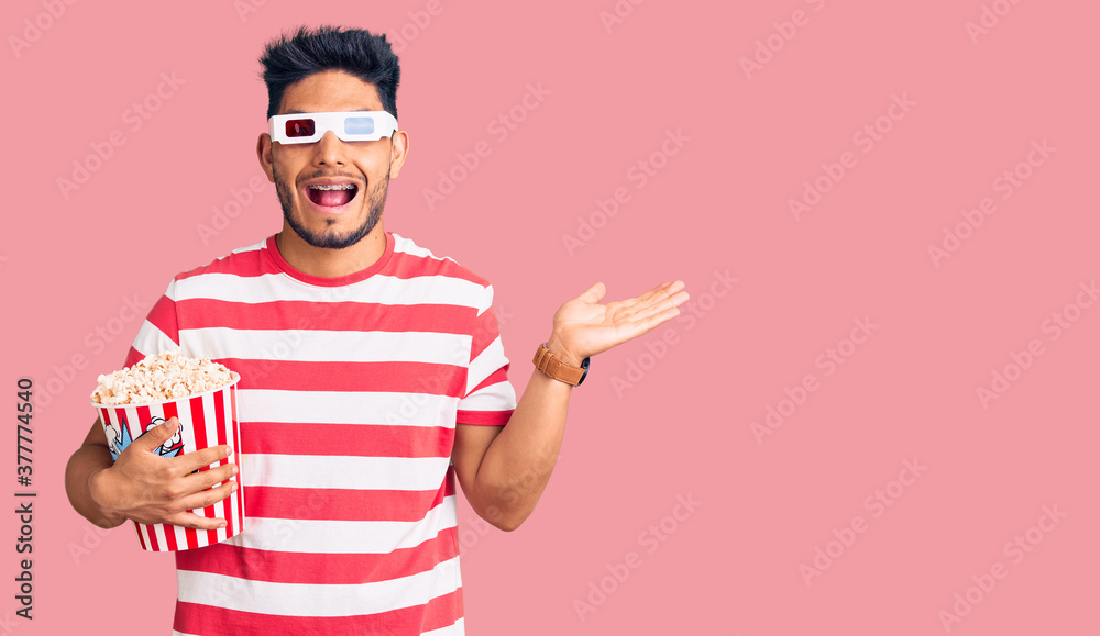 Handsome latin american young man wearing 3d glasses and eating popcorn at the movies celebrating victory with happy smile and winner expression with raised hands Handsome latin american young man wearing 3d glasses and eating popcorn at the movies celebrating victory with happy smile and winner expression with raised hands