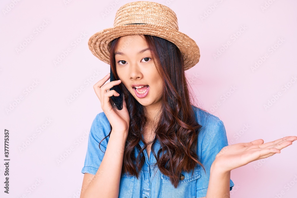 Young beautiful chinese girl wearing summer hat having conversation talking on the smartphone celebrating achievement with happy smile and winner expression with raised hand Young beautiful chinese girl wearing summer hat having conversation talking on the smartphone celebrating achievement with happy smile and winner expression with raised hand